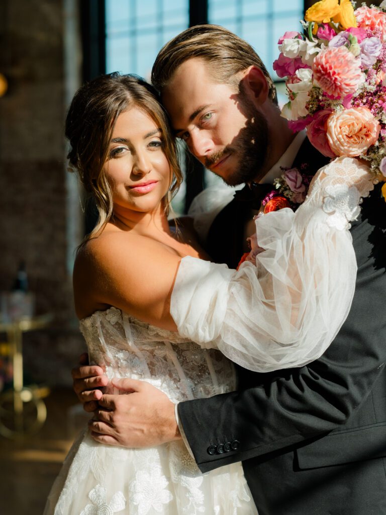 A bride and groom share an intimate embrace by a sunlit window, the bride in a lace off-the-shoulder gown with sheer sleeves, and the groom in a black suit holding a colorful bouquet of roses and dahlias.