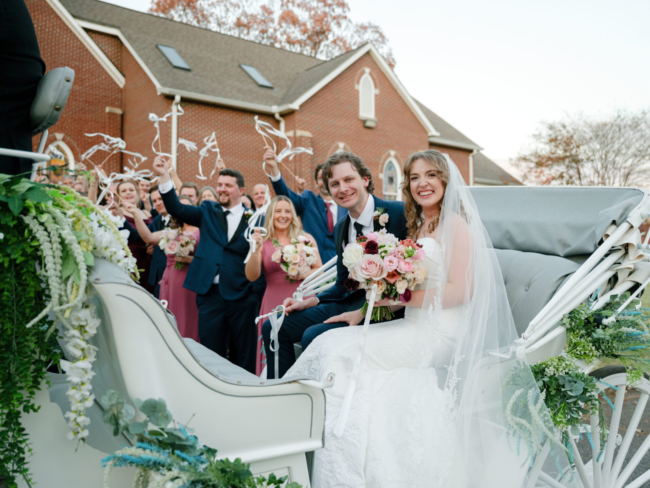 Bride and groom smiling in a white carriage surrounded by cheering guests waving ribbon wands outside a brick chapel